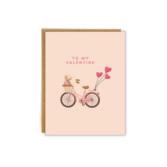 pink card with illustration of a bike, with flowers and a gift in the front basket. Trailing the bike is a bundle of heart balloons. Phrase "To my Valentine"